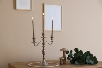 Stylish candlestick with burning candles and other decor on wooden table near beige wall indoors