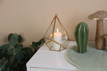 Burning candle in holder and other decor on cabinet near beige wall indoors