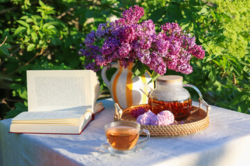 Beautiful lilac flowers in vase, tea, zephyrs and book on table in garden