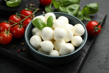 Tasty mozzarella cheese balls in bowl, basil, peppercorns and tomatoes on black table, closeup