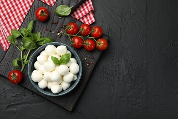 Tasty mozzarella cheese balls in bowl, basil, peppercorns and tomatoes on black table, top view. Space for text