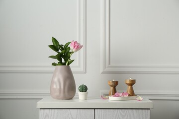 Elegant candlesticks with burning candles, tulip in vase and other decor on dresser near white wall