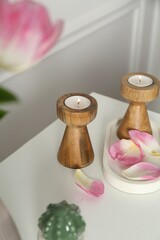Elegant candlesticks with burning candles and flower petals on dresser near white wall