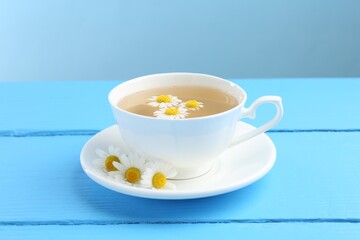 Delicious chamomile tea in cup and fresh flowers on light blue wooden table, closeup