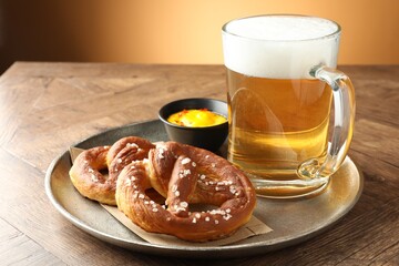 Tasty pretzels, sauce and glass mug beer on wooden table against beige gradient background, closeup