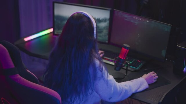 back view of female gamer wearing white headset and fluffy sweater playing immersive video game on dual monitors with rgb lighting, mechanical keyboard, and red microphone in dark gaming room setup