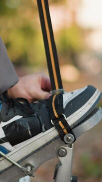 Close-up of person fastening shoes to spring stilts using orange and black straps, wearing gray trousers and long-sleeved shirt outdoors, blurred background, focus on adjustment process