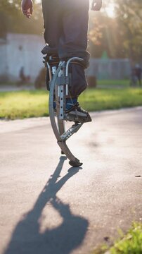 Guy walking on spring-loaded prosthetic legs outdoors in sunny park setting with greenery and paved path, wearing dark pants. Shadows are cast on path, capturing motion