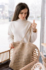 Stylish woman with red nails holding eco woven bag in cozy modern cafe with natural decor.