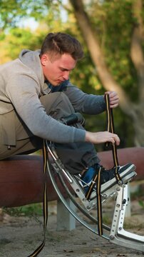 Young man with light brown hair seated on reddish-brown log in park, wearing light gray sweatshirt and khaki pants, focused on fastening spring stilts with concentrated or mildly frustrated expression