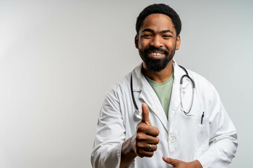 Smiling Black doctor giving thumbs up, isolated on neutral background. Young medical professional...