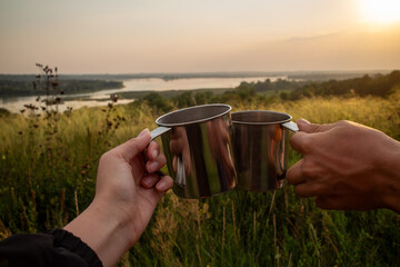 Two hands holding stainless steel camping mugs in a toast against a scenic sunset landscape with rolling hills and a winding river symbolizing outdoor adventure travel and shared moments in nature.