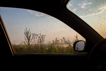 Looking out from inside a car through a window covered in raindrops reveals a hazy landscape of vegetation and a body of water under a soft evening light suggesting a journey on a wet road.