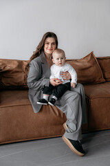 Stylish young mother with long dark hair in a gray stylish suit with her son in her arms sitting on the sofa. Handsome son with mom