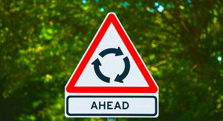 A roundabout ahead sign with a triangular shape and green trees in the background on a sunny day
