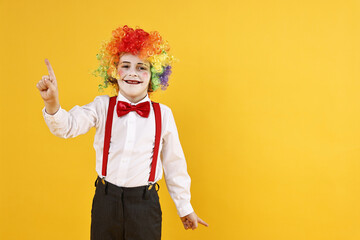 Emotional little boy dressed like clown pointing upwards on yellow background, space for text. Surprise party