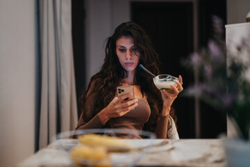 A woman sits at a table, focusing on her smart phone while holding a bowl of yogurt. The scene...