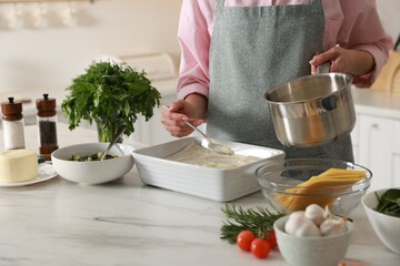 Woman spreading bechamel sauce onto spinach lasagna at marble table indoors, closeup