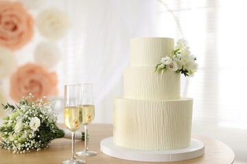 Delicious wedding cake with floral decor, glasses of champagne and bouquet on wooden table indoors