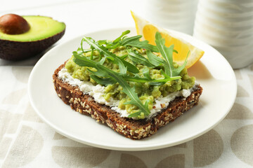 Delicious sandwich with avocado, cream cheese, arugula and lemon wedge on table, closeup