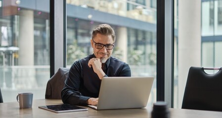 Plakat Business portrait - businessman using laptop computer in office, thinking. Happy middle aged man, entrepreneur working online.