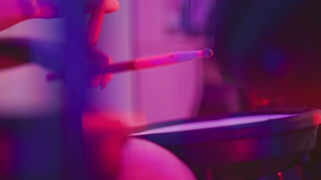 Close up of person holding drumsticks practicing on electronic drum in colorful studio illuminated with deep pink and purple lights emphasizing hand motion and musical intensity during rehearsal