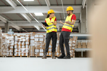 Two warehouse workers engage in a lively discussion while using a tablet in a bustling storage environment filled with boxes.
