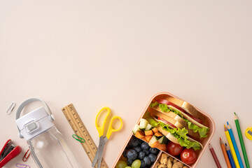 Lunchbox with a sandwich, vegetables, and fruits beside school supplies on a desk