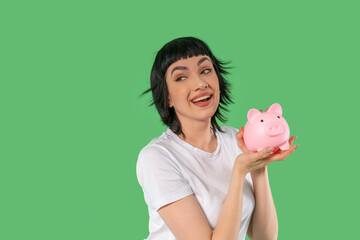 Smiling young woman holding a pink piggy bank against a vibrant green background while promoting...