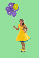 Young woman in yellow dress holds balloons and a small dog while balancing on a platform against a green background