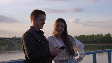 Young couple using smartphone near river at sunset, sharing social media content
