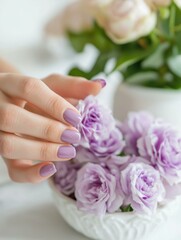 Close-up of a woman's hand with a manicure. the hand is resting on a white surface with a small white bowl filled with purple roses. the roses are in full bloom and have delicate petals.