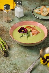 Fresh asparagus puree soup with asparagus pieces and fried bacon in a pink ceramic plate on a green concrete background. Recipes asparagus.