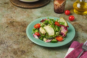 Fresh vegetable salad with fried asparagus, arugula, cucumber and cherry tomatoes on a green plate on a green concrete background. Vegetable salads, vegan recipes. Seasonal vegetables.