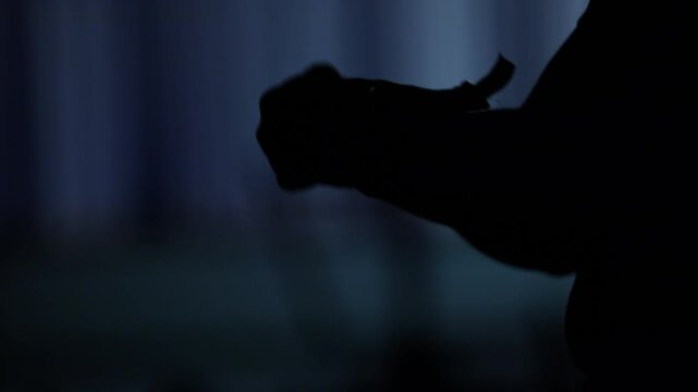 Silhouette of hands in low light, first manipulating a thin object then clasping together in prayer. Mysterious, ritualistic atmosphere with dark blurred background evokes contemplation.