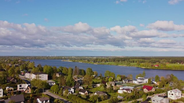 Slow drone flyover above Katlakalns residential area showing houses, tree lines, Daugava river shoreline, and Riga skyline on horizon under scattered clouds and afternoon light, springtime