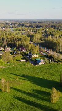 Vertical drone gliding above Katlakalns countryside shows green fields, scattered houses, Daugmale road, forest line, distant Riga skyline under warm sunset light, slow motion