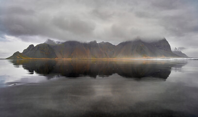 Calm water reflecting dramatic Vestrahorn mountain range under cloudy sky