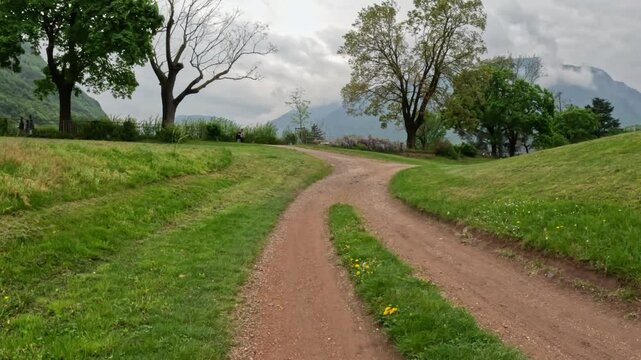 OV walk on a forked dirt track through green riverbank meadows, framed by mature trees and misty Mendola foothills, leading from Bolzano&rsquo;s Talvera promenade toward the historic Castel Roncolo
