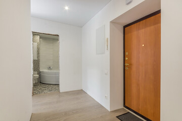 An empty apartment hall with a brown front door. Laminated floor. View of the bathroom in the doorway without doors and unfinished repairs.