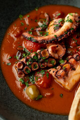 Savor a beautifully presented seafood dish with tender octopus pieces immersed in a vibrant tomato sauce, garnished with fresh herbs, at a cozy restaurant while enjoying an evening meal
