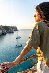 Woman standing on a balcony overlooking a tranquil ocean scene with sailboats, enjoying the serene sunset and natural beauty