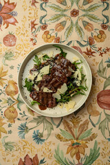 A beautifully plated dish featuring tender slices of beef on a bed of fresh greens, garnished with shavings of cheese, all set against an artistic floral tablecloth in a charming eatery
