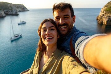 Happy couple taking a selfie with a stunning ocean backdrop featuring sailboats and cliffs, capturing a memorable travel moment