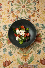 A beautifully arranged salad featuring fresh greens and strawberries topped with creamy cheese, elegantly presented on a decorative tablecloth. The ambiance is inviting and sophisticated