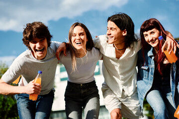 Four cheerful young people are walking together outdoors, enjoying a sunny day and holding...