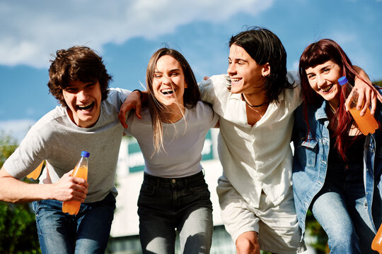 Four cheerful young people are walking together outdoors, enjoying a sunny day and holding orange-colored drinks