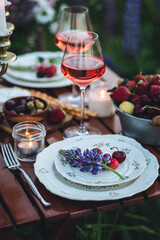 Beautiful elegant romantic dinner table decor, surprise day arrangement. Marriage proposal, engagement private party for two in a meadow. Sunset, golden hour. Cheese board, rose wine, fruits, lupine