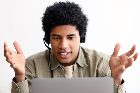 Web based education concept. Happy black teen student in headset talking to his college tutor online on laptop, indoors. Handsome teenager taking part in web conference or internet lesson from home