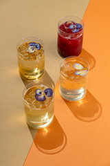 Four elegant glasses filled with refreshing summer drinks sit on a vibrant orange and beige surface. Each drink is garnished with colorful edible flowers, adding a touch of beauty